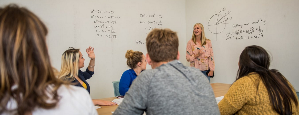 Students watching an instructor write equations on a white board.