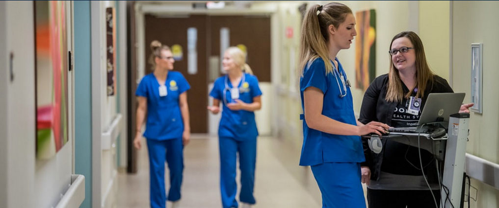 Nurse talking to a student next to a computer. Two other nursing students are walking down the hallway.