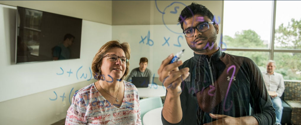 Student writing equations on a board with professor standing next to them.