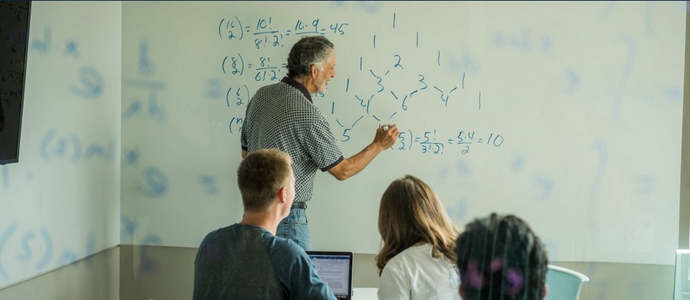 Instructor writing equations on a white board with students watching.