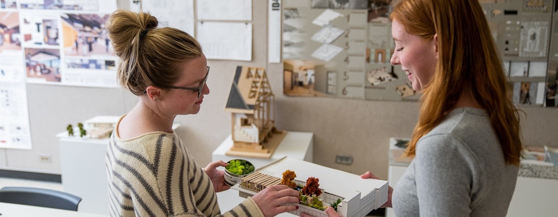 Alt text: Two students in a design studio examine a detailed architectural model featuring miniature trees and structures. One student holds the model while the other adds a piece to it. The background displays various architectural plans, posters, and scale models pinned to the walls, indicating a creative and academic environment.