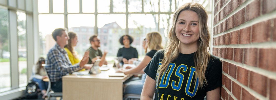 Student smiling for the picture with a group of students sitting behind her at a table talking.
