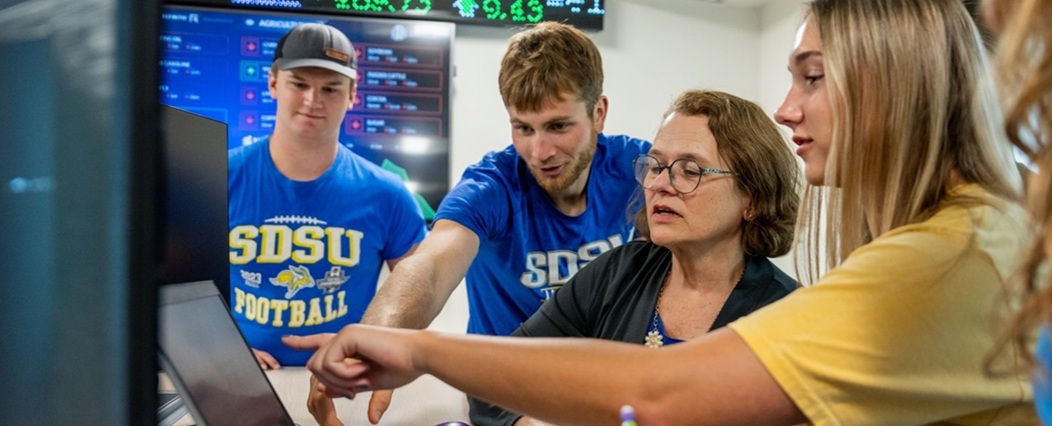 A group of students and professor collaborate around computer monitors in the finance trading lab. Digital stock market ticker displays and data screens are visible in the background.