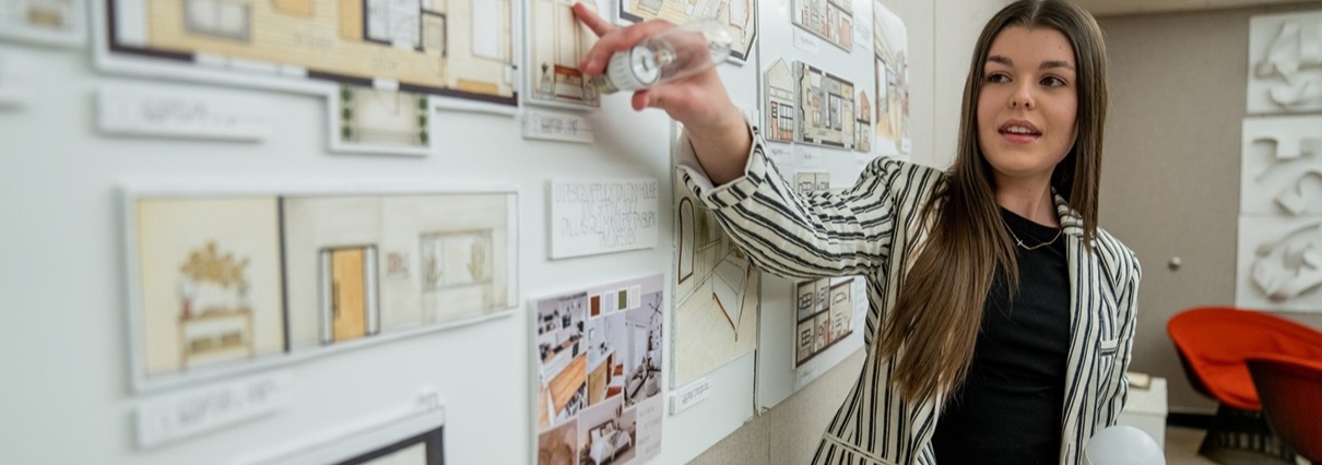 A student presents an interior design project, pointing to a display board filled with floor plans, renderings, and material samples.